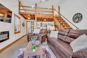 Ein geräumiges Wohnzimmer mit hohen Decken, freiliegenden Holzbalken, einem braunen Ledersofa, einem Couchtisch mit Dekoration und einer Treppe, die zu einer oberen Ebene führt.