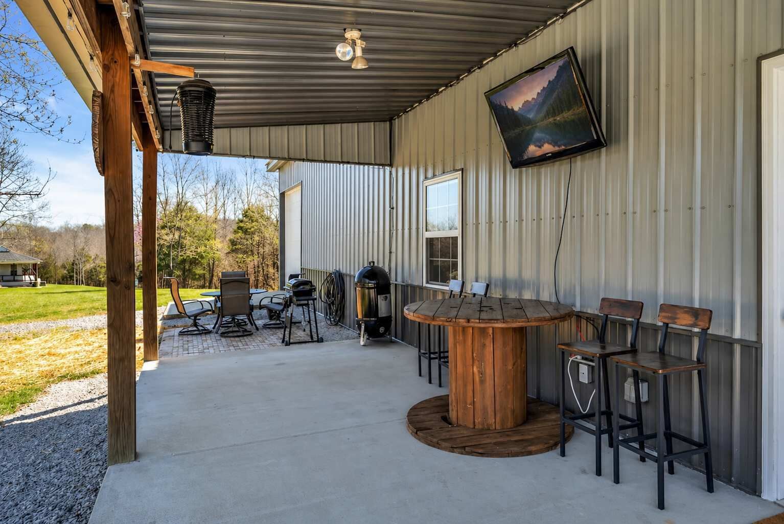 Un patio exterior con una mesa redonda de madera y sillas, una parrilla y un televisor montado en la pared.