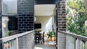 Een balkon met een zwarte bakstenen muur, een houten leuning, een eethoek met tafel en stoelen, en een potplant.