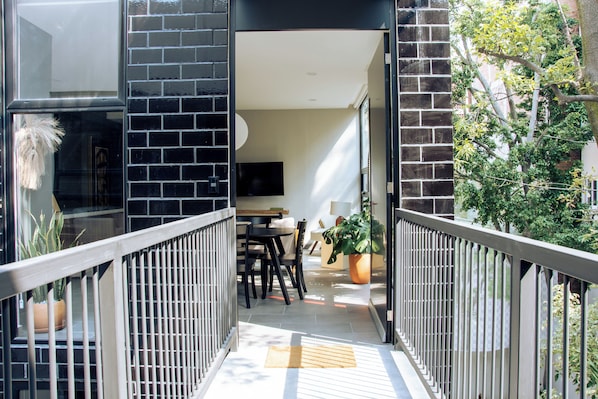 A balcony with a black brick wall, a wooden railing, a dining area with a table and chairs, and a potted plant.