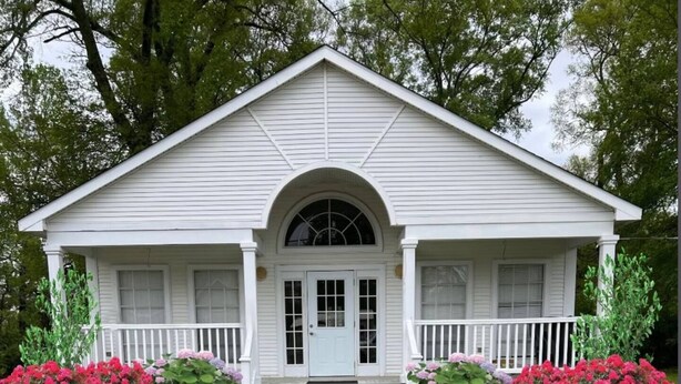 Une maison blanche à deux étages, dotée d’un porche à l’avant, d’une entrée centrale en arc de cercle et d’un petit jardin devant, agrémenté de fleurs roses et rouges.