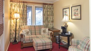 A cozy room with a plaid sofa set, a coffee table, and a lamp. There are two windows with floral curtains, and a framed picture on the wall. - Small Private suite with Electric Fireplace and Pet Friendly (Blowing Rock)