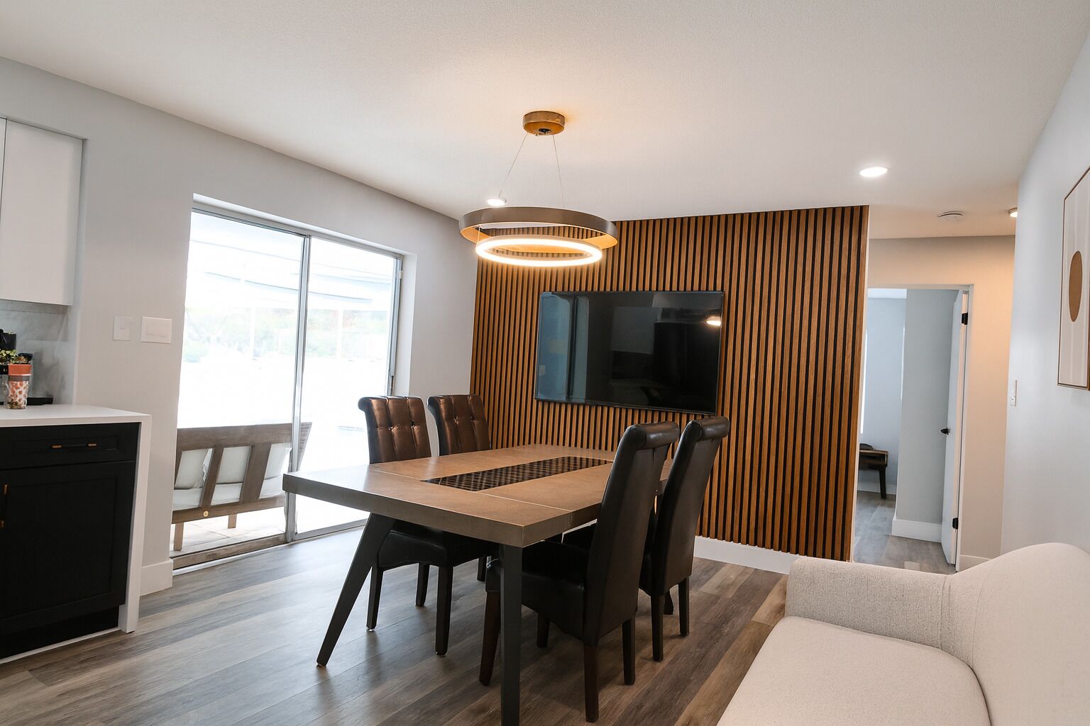 A modern dining area with a wooden table, black chairs, and a television mounted on a wooden panel wall.