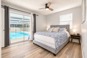 Une chambre avec un lit, une table de chevet, un ventilateur de plafond, une fenêtre avec des stores et une vue sur une piscine extérieure.