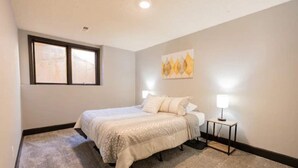 Quarto com cama, mesa de cabeceira, luminária, janela e um quadro na parede.