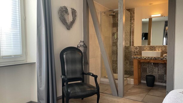 A hotel room with a wooden floor, a black leather chair, a heart-shaped decoration on the wall, a shower area, and a bathroom with a sink and mirror. - GITE VAUBAN NEUF-BRISACH (NEUF BRISACH)