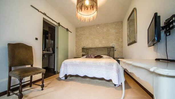 A bedroom with a bed, a chair, a desk, a television, and a stone wall. - Saint Martin Guesthouse LE Vauban (NEUF BRISACH)