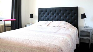 Een net opgemaakt bed met een gestoffeerd hoofdeinde, een nachtkastje met een lamp en een rode kruk.