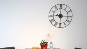A small table with two black chairs, a wall clock, and a few items on the table including a yellow bowl, a red pot with a plant, and a water bottle. - Briancon Guesthouse LE Vauban (NEUF BRISACH)