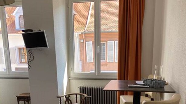 A small, well-lit room with a bed, a desk, a chair, and a radiator. - Camaret Guesthouse LE Vauban (NEUF BRISACH)