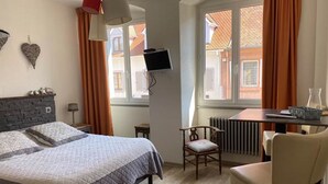 A bedroom with a bed, a desk, a chair, a TV, and a window with curtains. - Camaret Guesthouse LE Vauban (NEUF BRISACH)