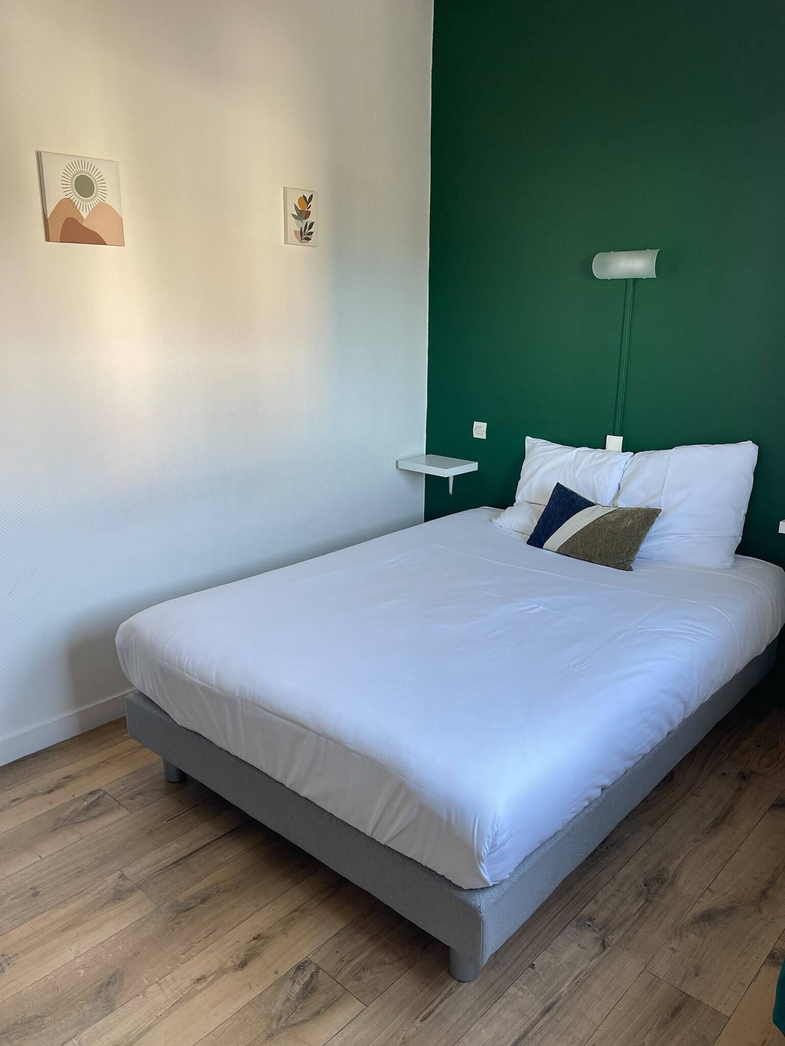 A neatly made bed with white bedding, a green accent wall, and a small bedside table with a lamp.