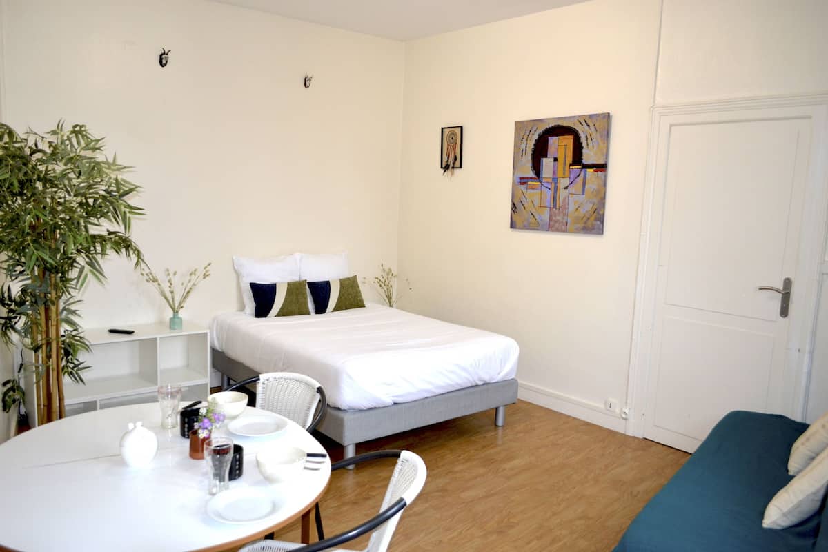 A bedroom with a bed, a bedside table, a chair, a dining table with chairs, and a potted plant.