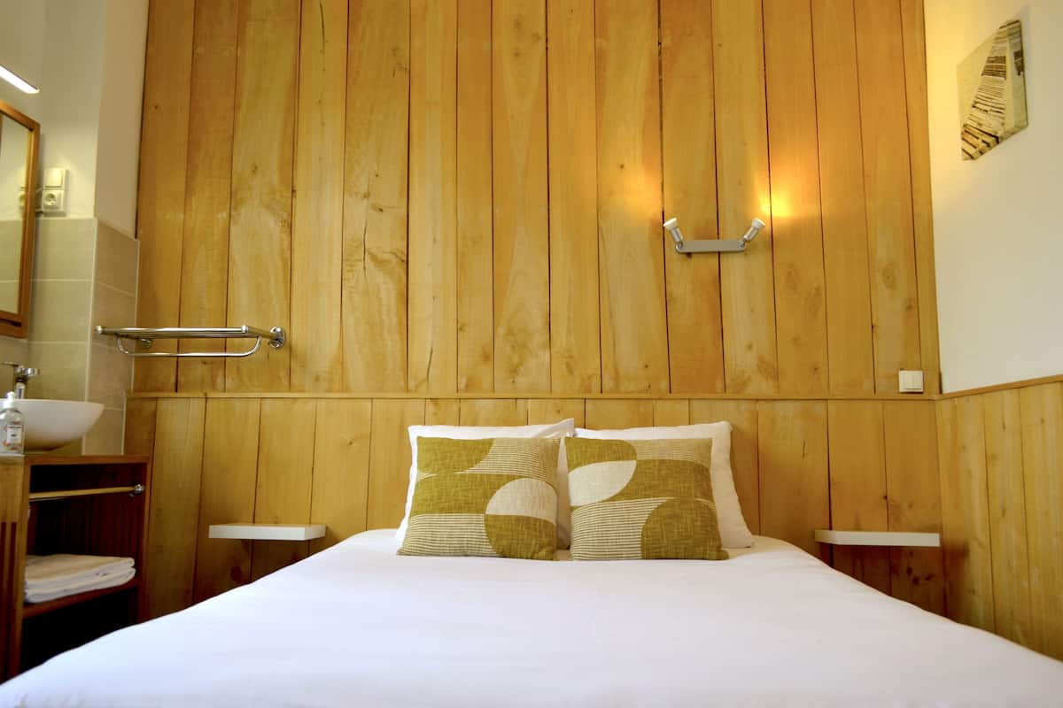 A hotel room with a wooden headboard, a bed with white bedding, two pillows with a geometric pattern, a bedside table, a nightstand, a towel rack, and a mirror.