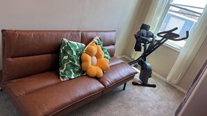 Ein braunes Ledersofa mit einem Kissen mit grünem Blattmuster und einem gelben Strickkissen. Ein Heimtrainer steht an einer Wand neben einem Fenster.