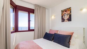 Un dormitorio con cama, ventana con cortinas, un cuadro en la pared y vista al mar.