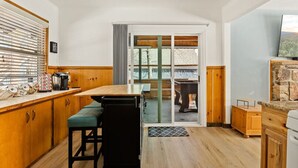 Une cuisine compacte avec des meubles en bois, un coin repas avec un banc, une porte-fenêtre coulissante donnant sur une table de billard et une cheminée en pierre.