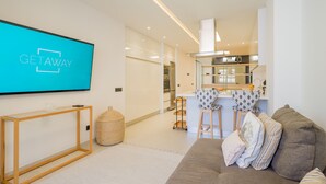 A modern living room with a grey sofa, a TV mounted on the wall, and a kitchen area in the background.