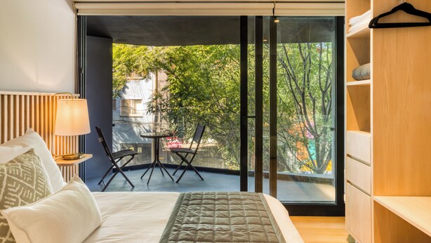 Een moderne slaapkamer met een groot bed, een nachtkastje met een lamp, een stoel en uitzicht op een balkon met een tafel en stoelen.