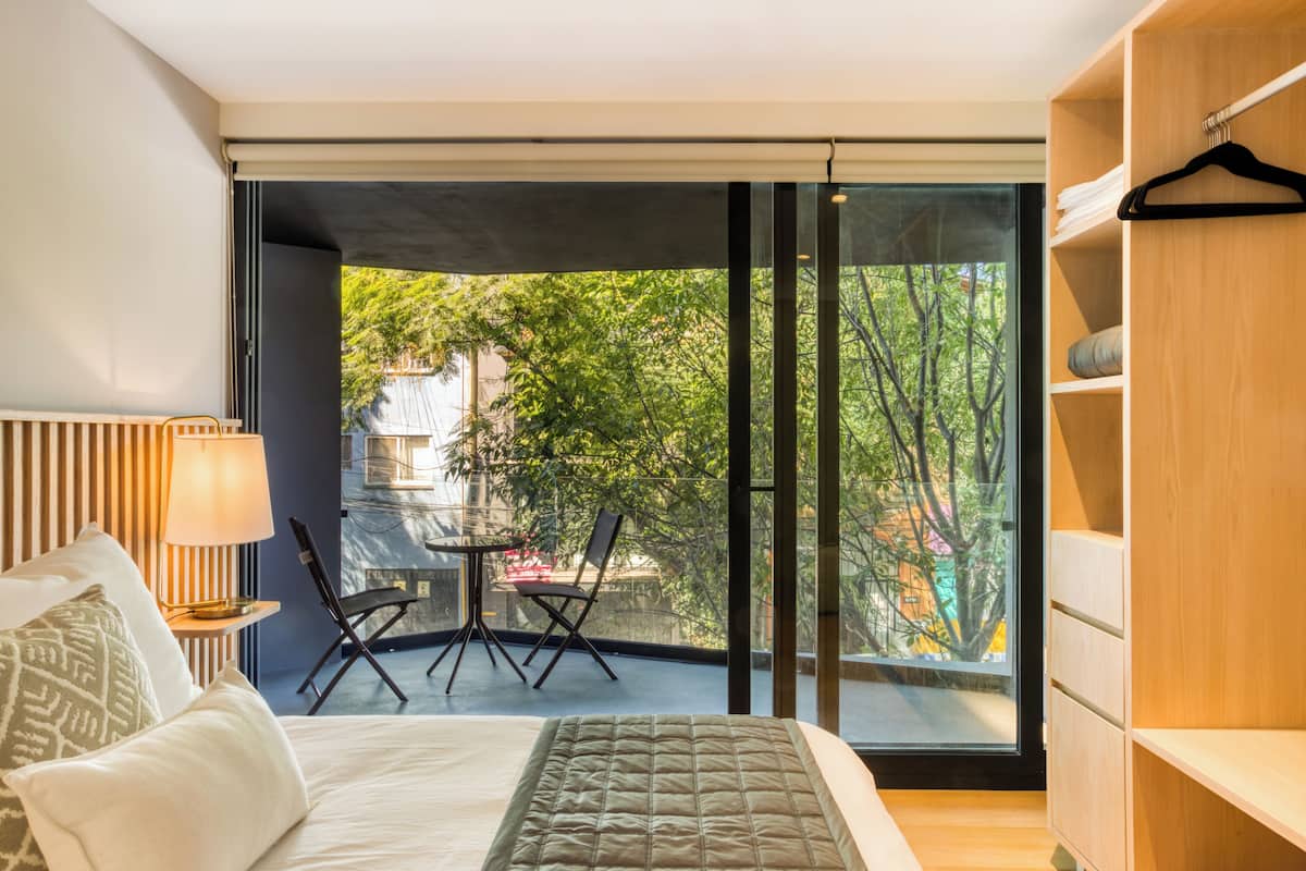 Ein modernes Schlafzimmer mit einem großen Bett, einem Nachttisch mit Lampe, einem Sessel und Blick auf einen Balkon mit Tisch und Stühlen.