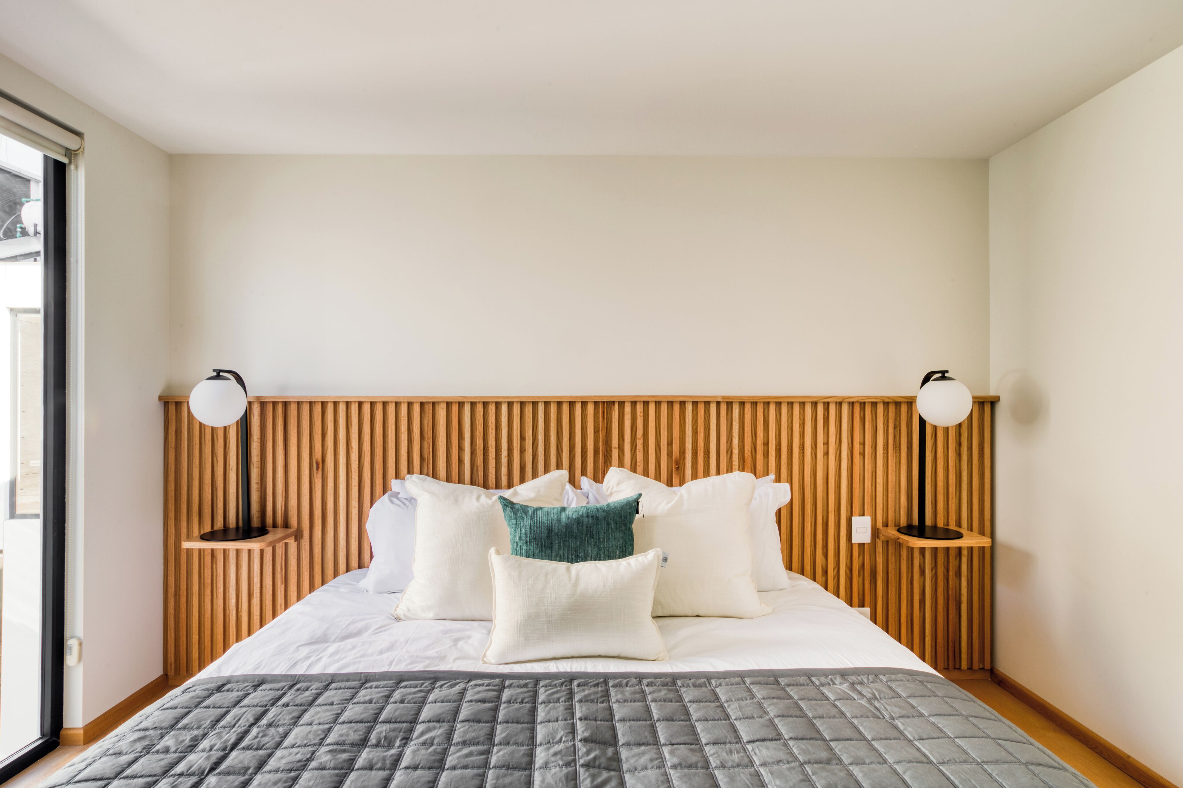A neatly made bed with a grey quilt, white pillows, and a green accent pillow. There are two bedside tables with lamps on either side of the bed. The room has wooden paneling on the wall behind the bed.