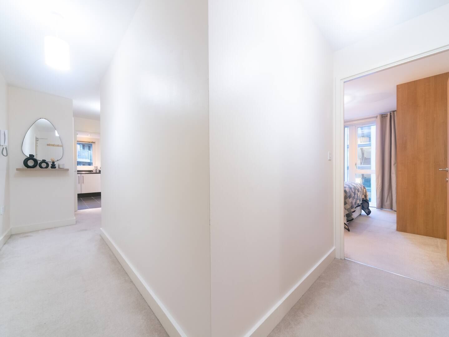Un corridoio che conduce a una camera da letto con una porta in legno, uno specchio con cornice decorativa e vista su un balcone con finestra.