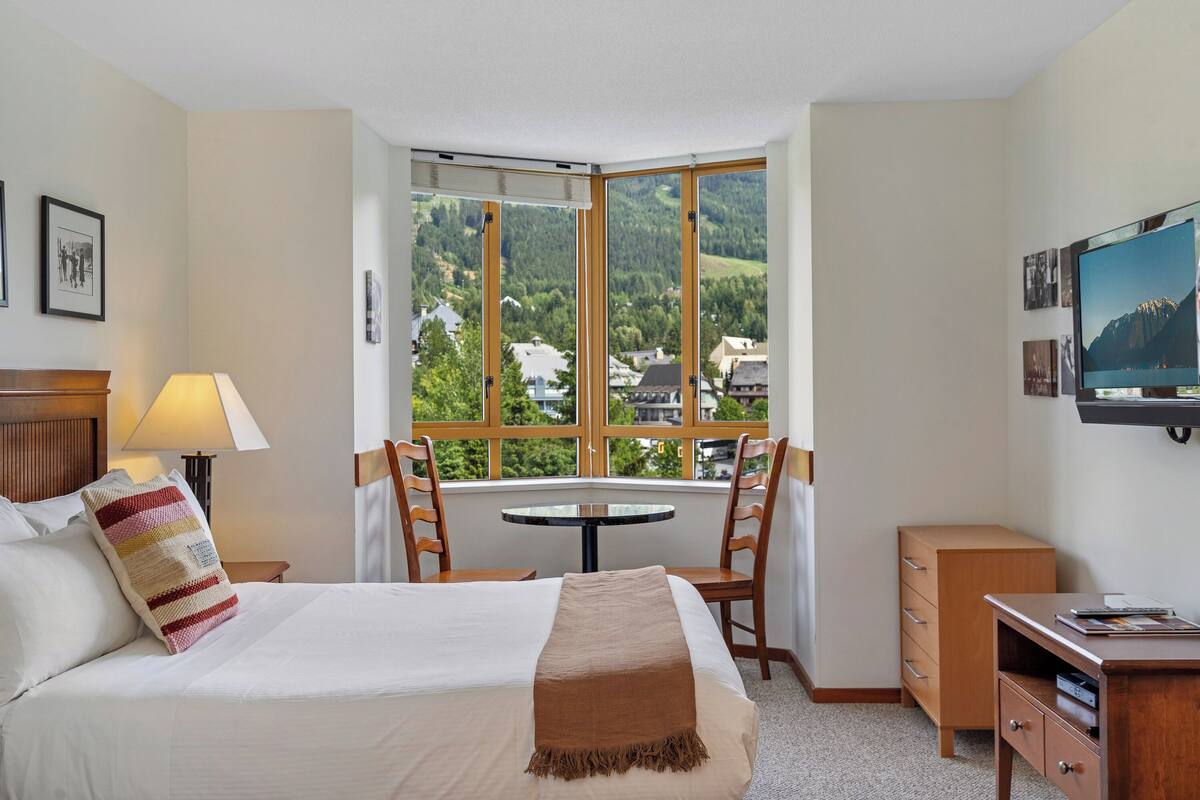 A hotel room with a bed, a desk, a chair, a table, and a TV.