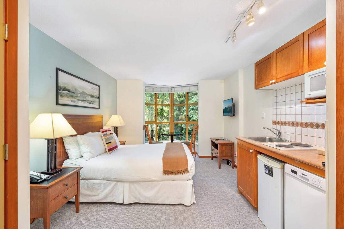 A hotel room with a bed, bedside lamps, a window with a view of greenery, a small table, a microwave, and a dishwasher.