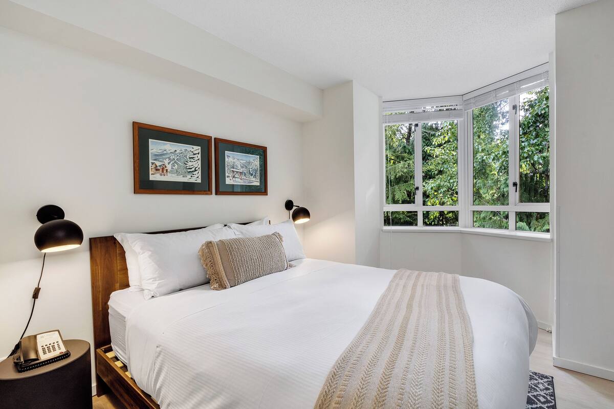A bedroom with a large bed, two framed pictures on the wall, a bedside lamp, and a window with a view of greenery.