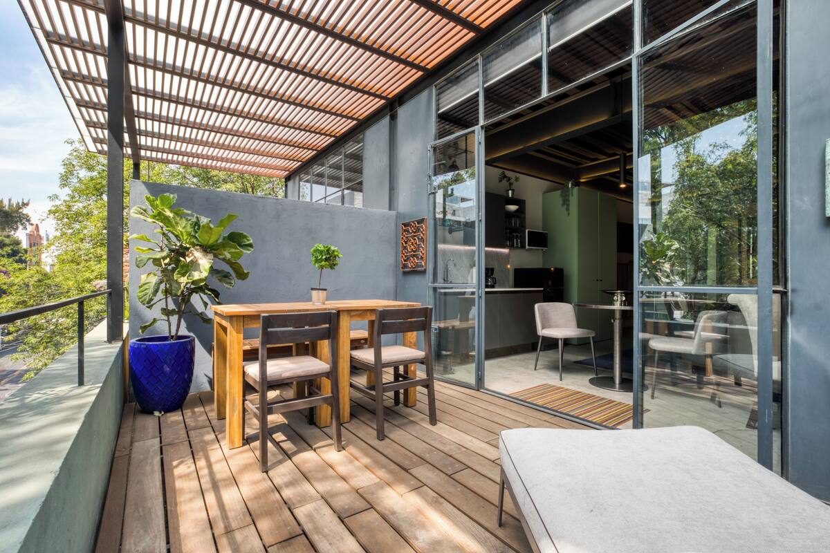 A modern outdoor deck with a wooden table and chairs, a large potted plant, and a glass wall leading to an indoor area with seating.