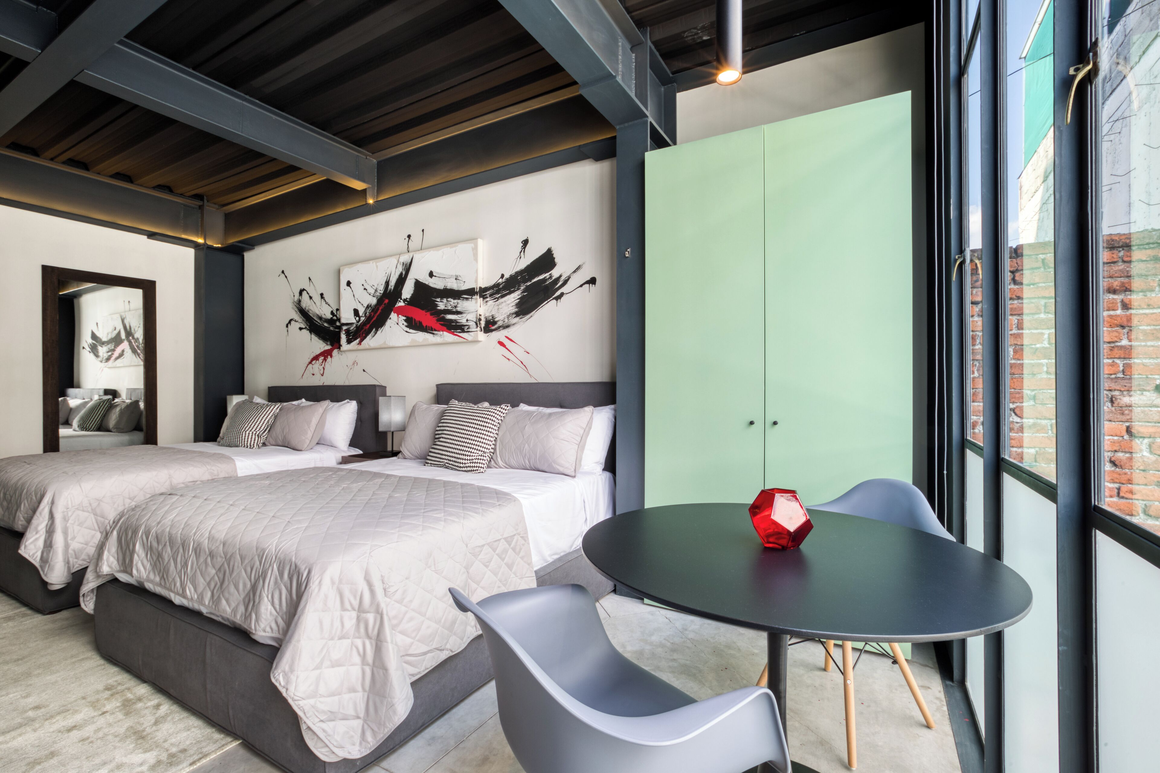 A modern bedroom with two beds, a large abstract painting, a green wardrobe, and a round table with a red object.