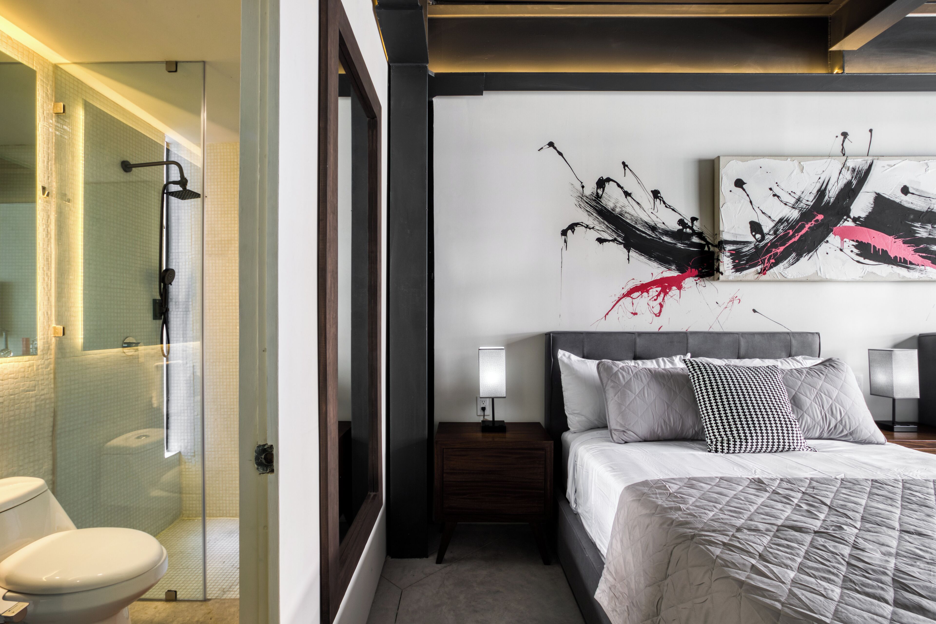 A modern hotel room with a glass-enclosed shower, a neatly made bed with a grey quilt, and a large abstract painting on the wall.