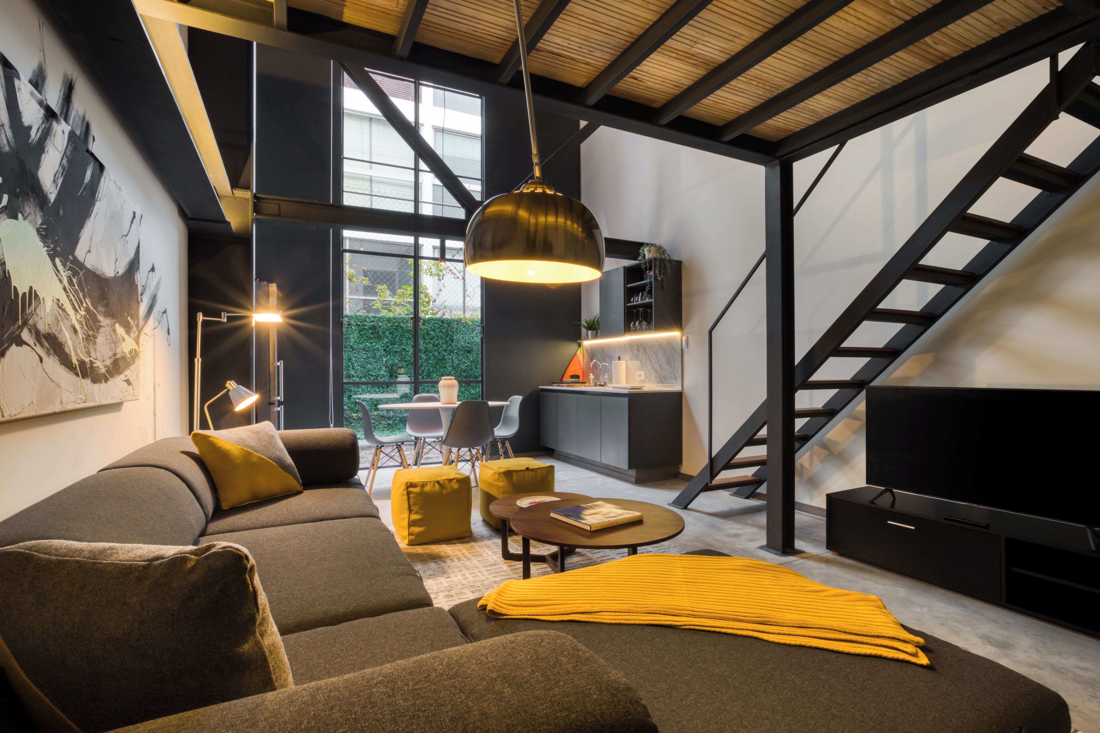 Ein modernes Wohnzimmer mit einer Couch, Poufs, einem Couchtisch und einem Fernseher. Eine Treppe führt auf eine höhere Ebene.