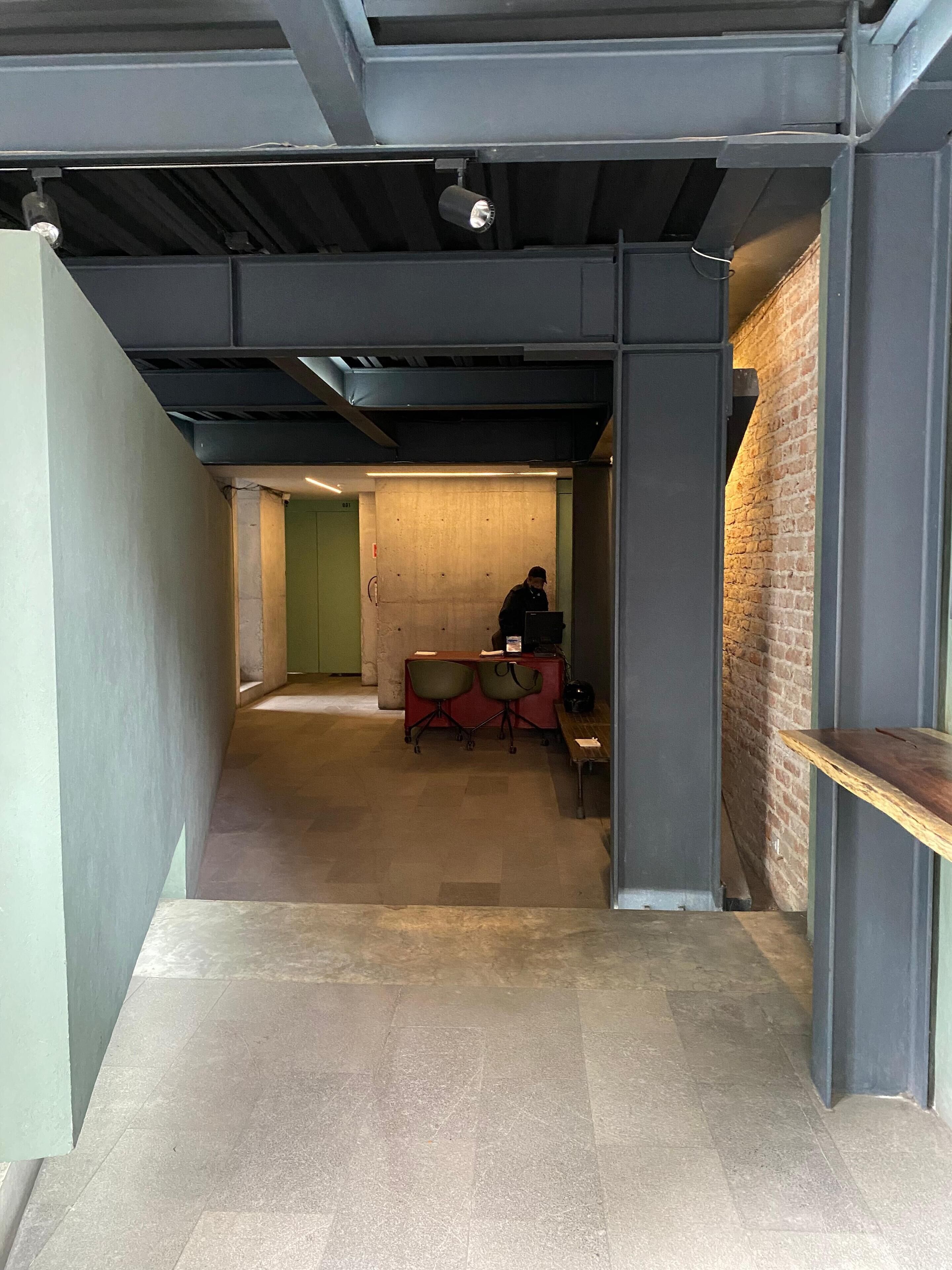 A hallway with a brick wall, a desk with chairs, and a person sitting at the desk.