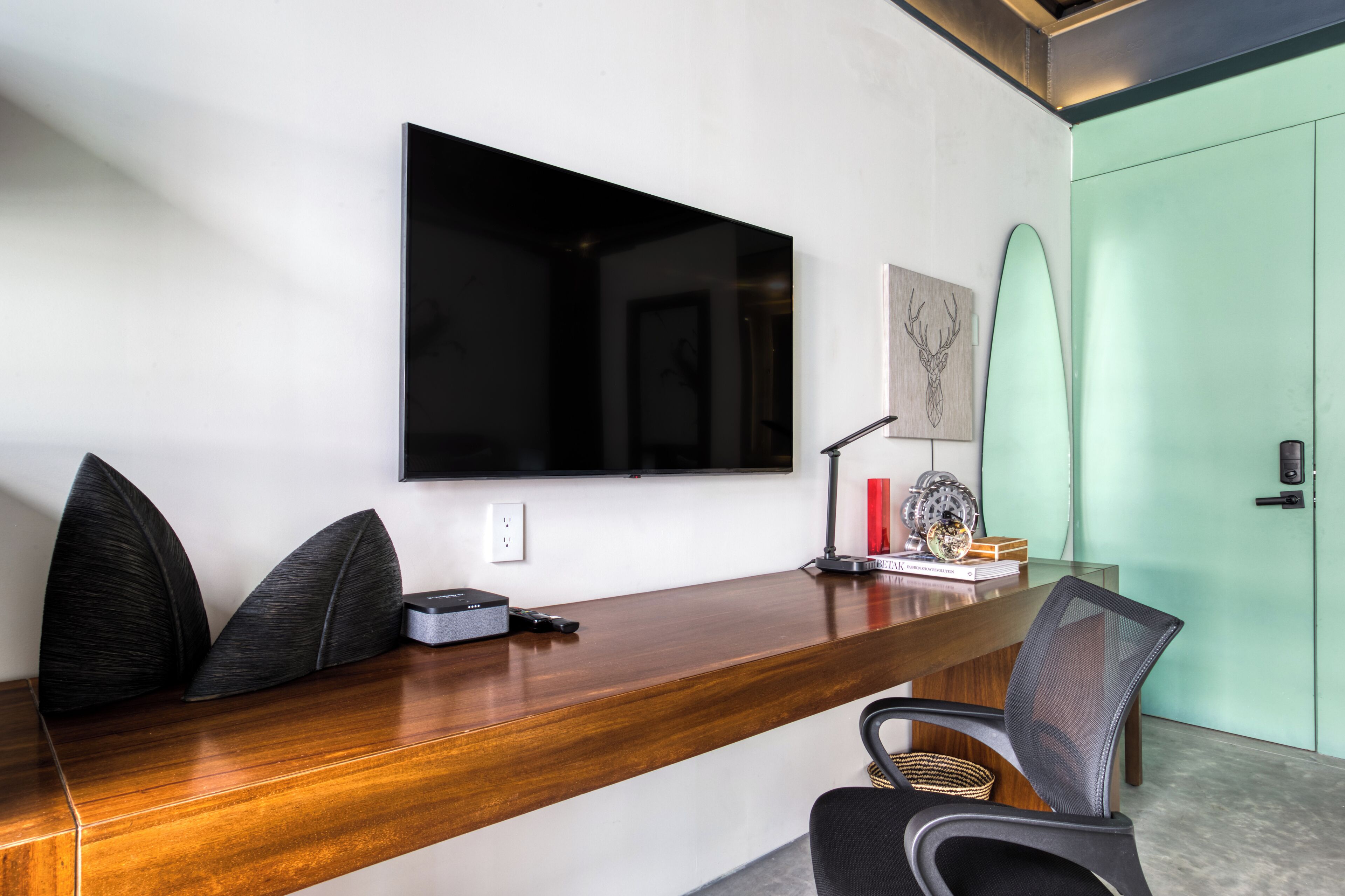 A modern office space with a large flat-screen TV mounted on the wall, a wooden desk with a chair, a surfboard, and a framed picture.