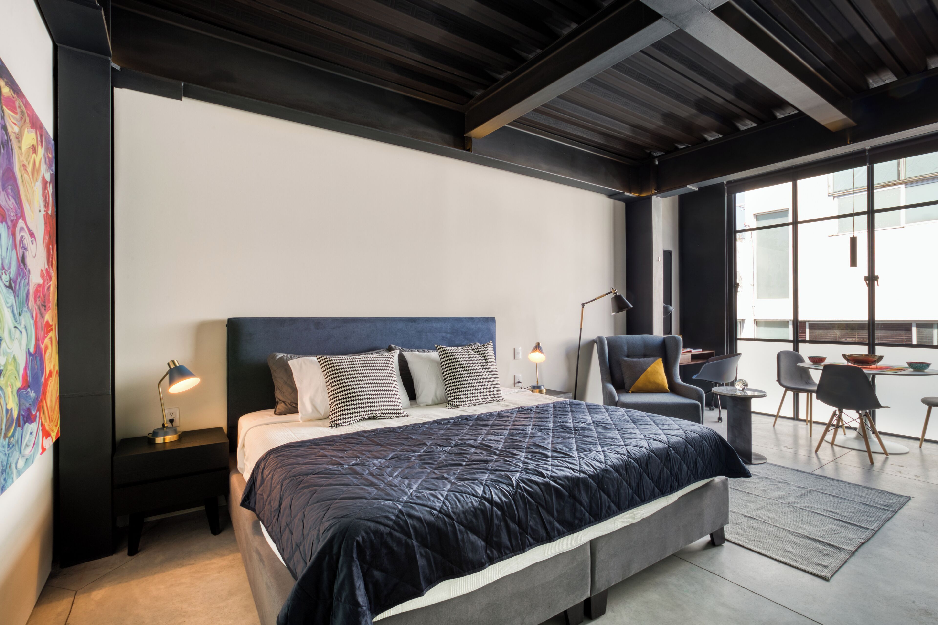 A modern bedroom with a large bed, a nightstand with a lamp, a chair, and a dining area with chairs and a table.