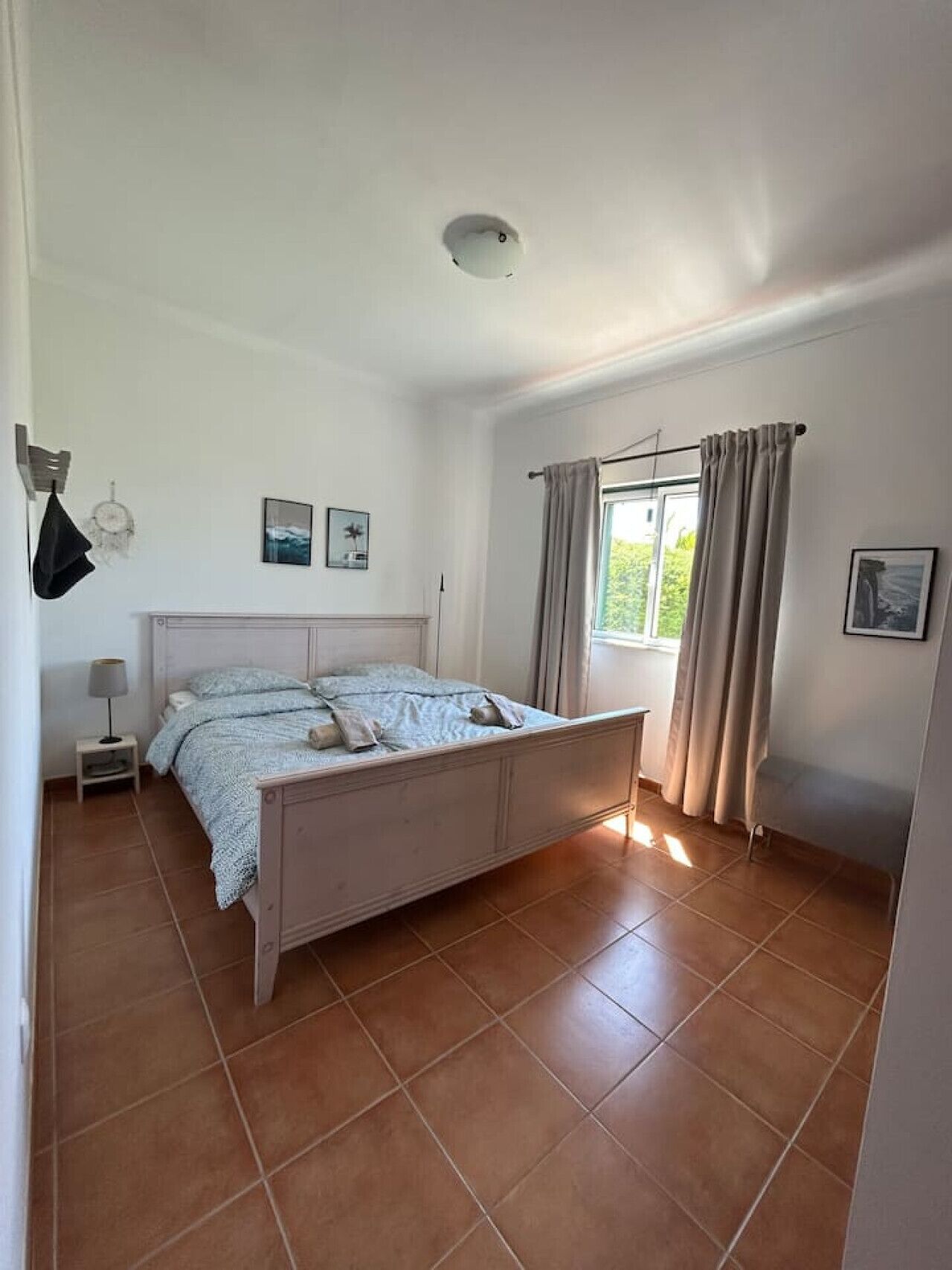 A bedroom with a bed, a window with curtains, a bedside table, and a wall-mounted light fixture.