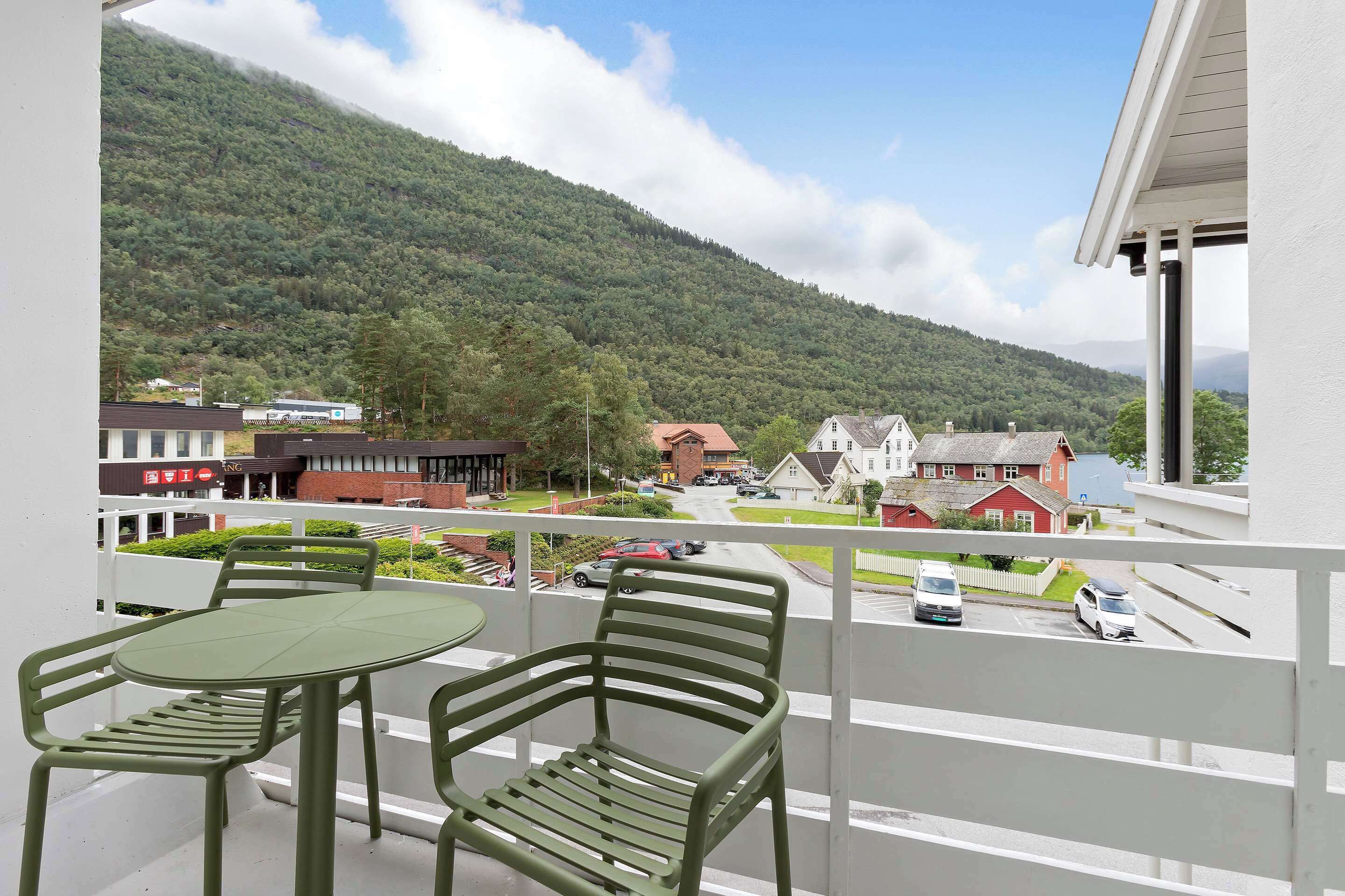 A balcony with two green chairs and a table, overlooking a street with cars and houses, and a mountain in the background.
