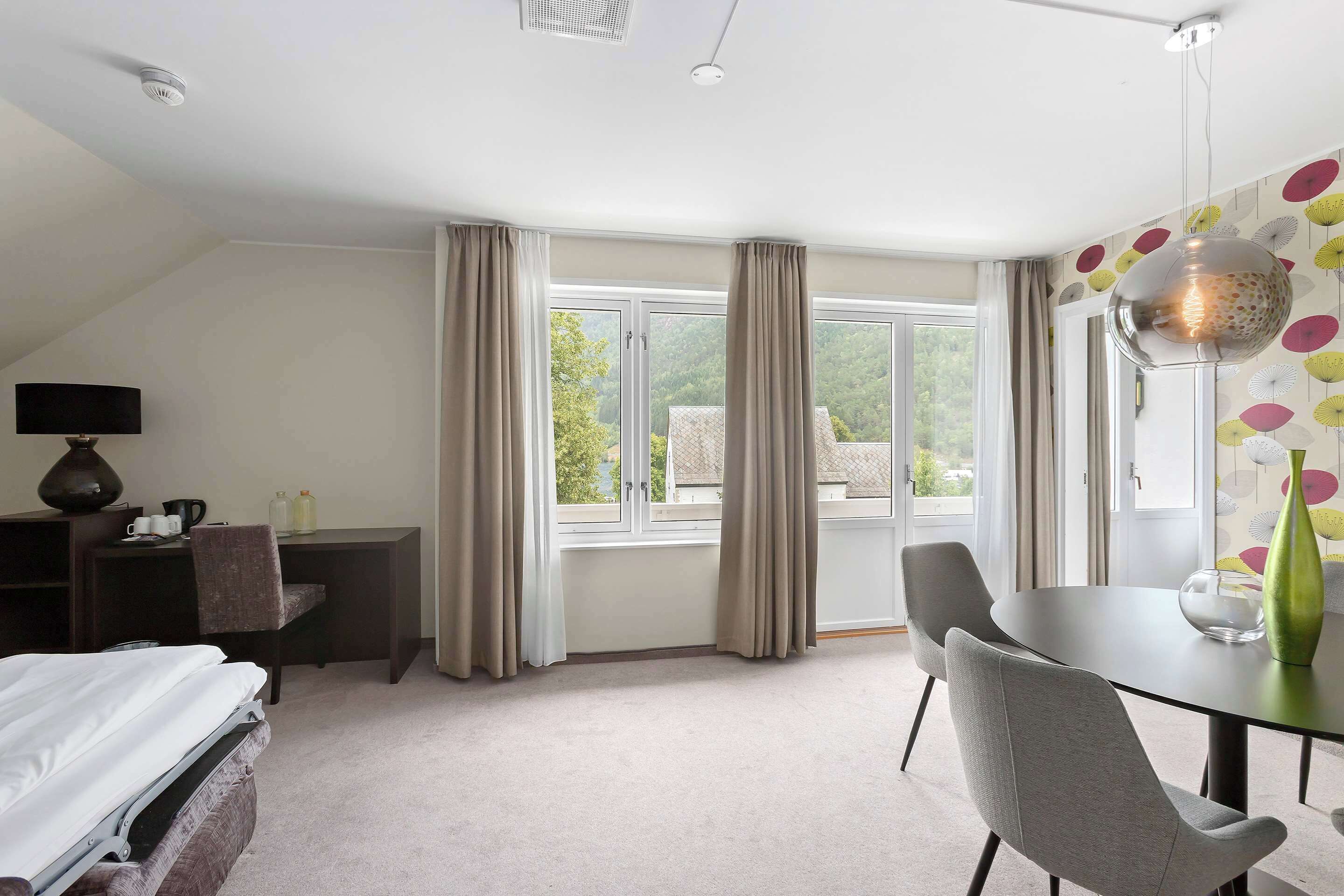 A hotel room with a bed, a desk with a chair, a dining table with chairs, and a large window with curtains.