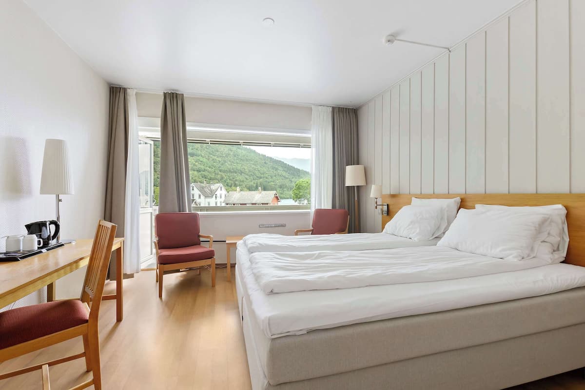 Una habitació d'hotel amb un llit gran, dues cadires, un escriptori amb una làmpada i vista a un turó verd.