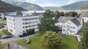 Ein weißes Gebäude mit vielen Fenstern und einer Treppe, umgeben von Grün und mit Blick auf einen See und Berge.