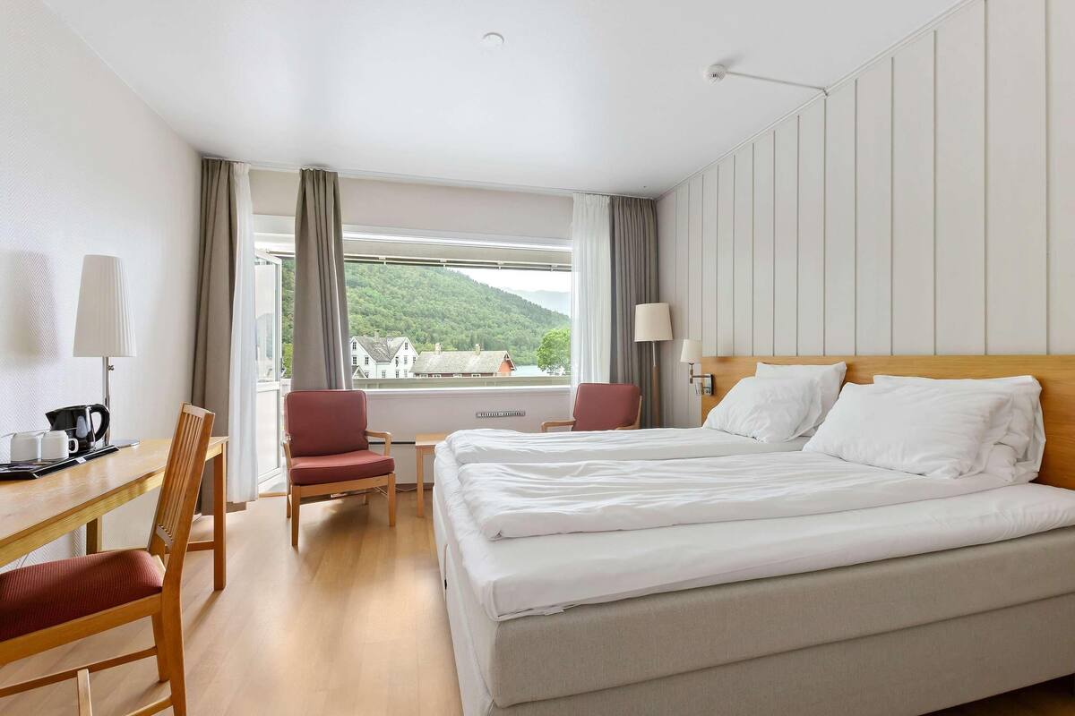 Habitació d'hotel amb un llit, dues cadires, un escriptori amb una làmpada i una finestra amb cortines.