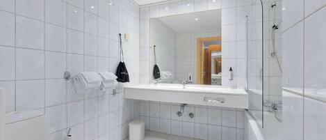 A modern bathroom with white tiles, a large mirror, a sink, a toilet, and a shower.