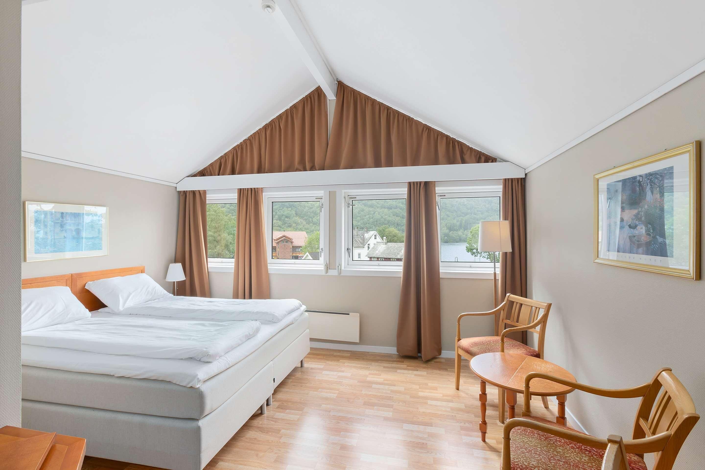 A hotel room with a bed, a desk, two chairs, a window with curtains, and a painting on the wall.