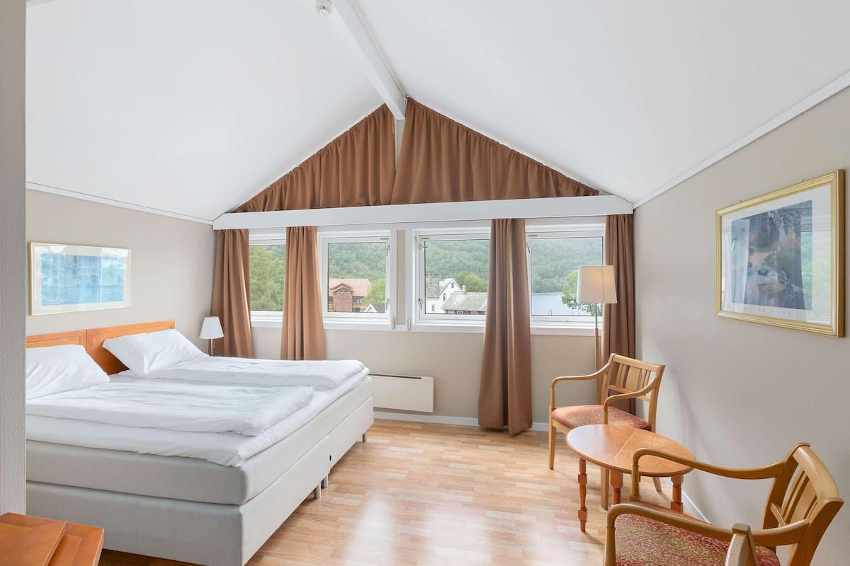 Una habitació d'hotel amb un llit, una taula de treball, dues cadires, una finestra amb cortines i un quadre a la paret.