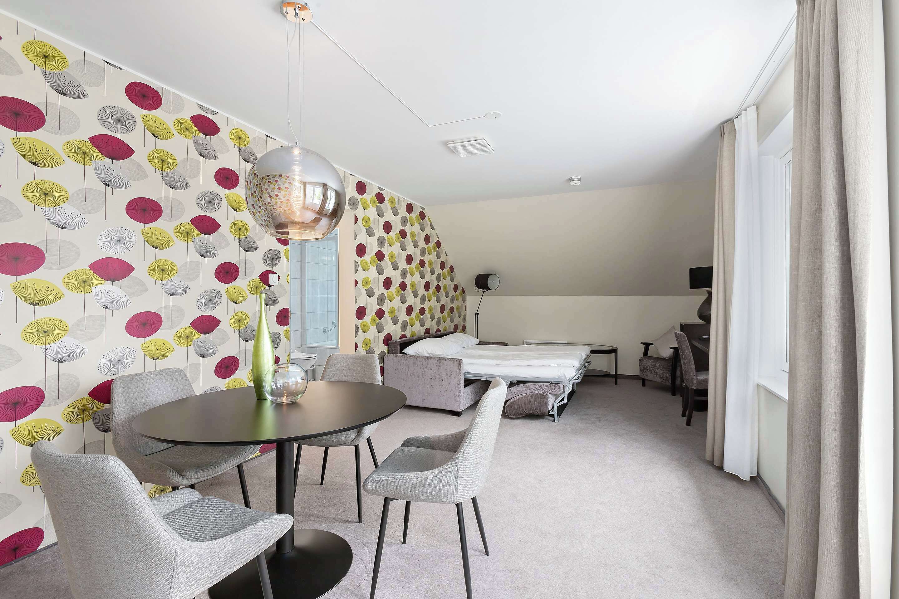 A modern hotel room with a polka dot wallpaper, a round table with chairs, a bed, a desk, and a chair.