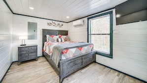 A bedroom with a bed, a nightstand, a dresser, a wall-mounted TV, and a window with blinds. - Broken Bow Cabin w/ Hot Tub, Fire Pit & Creek View (Broken Bow)