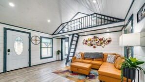 A spacious living room with a large sectional sofa, a wooden floor, a staircase, and a decorative wall piece. - Broken Bow Cabin w/ Hot Tub, Fire Pit & Creek View (Broken Bow)