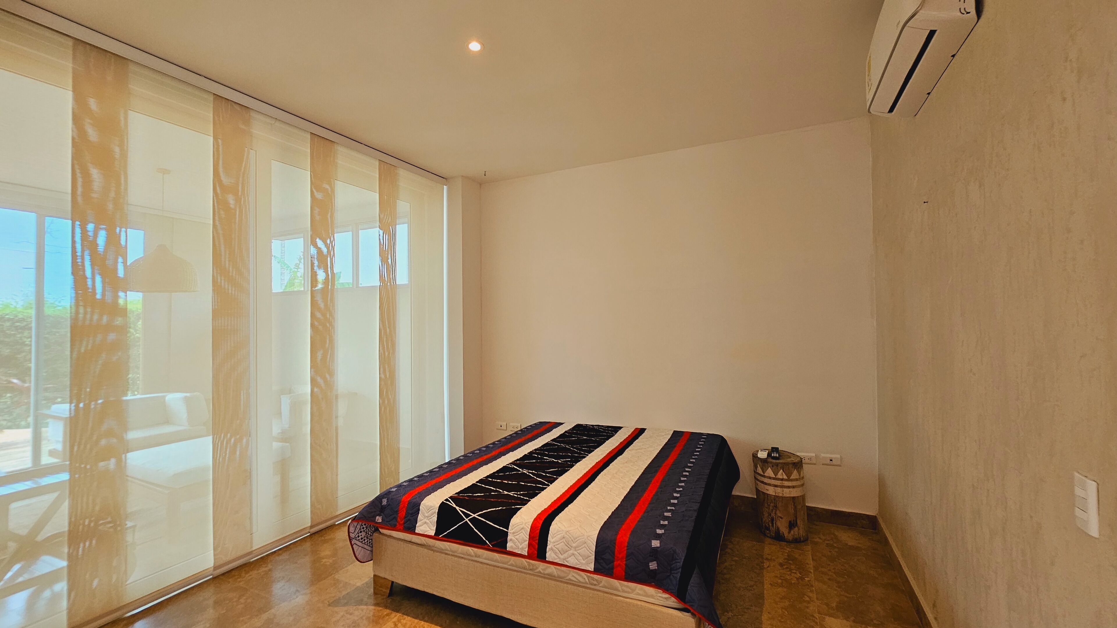 Une chambre à coucher avec un lit, une fenêtre avec des stores, un appareil de climatisation et un petit tabouret en bois.