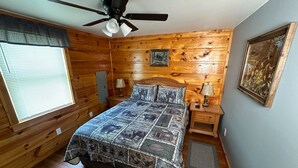 Un dormitorio con piso de madera, una cama con colcha estampada, un ventilador de techo, dos mesitas de noche con lámparas, una ventana con persianas y un cuadro en la pared.